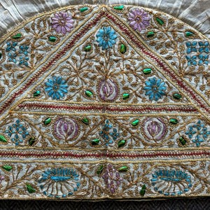 May include: Cream-coloured textile with detailed embroidery. It has a semi-circular shape, showcasing floral and geometric patterns in blue, purple, and green, all outlined with gold thread. The edge is ruffled.