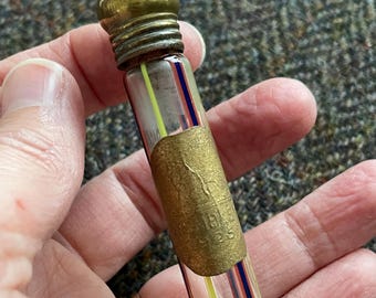 May include: A small, antique glass vial with a brass cap and a decorative label. The vial features colorful stripes of red, blue, and yellow. The label has embossed text. A vintage collectible.