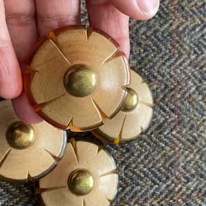 May include: Four wooden flower-shaped buttons with brass centers and amber-colored edges. One button is held by a hand, with the others scattered on a textured, multi-colored fabric background. The buttons have a unique, carved design.