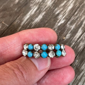 May include: A double row of turquoise and clear rhinestones set in a gold tone metal. The stones are round and have a faceted finish.