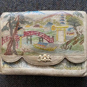 May include: A white leather clutch with a scalloped edge and a detailed embroidered scene of a Japanese village with a bridge, pagoda, and trees. The clutch has a decorative ivory-colored clasp with three figures.