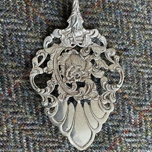 May include: A silver serving spoon with an ornate, floral design. The spoon has a long, slender handle and a wide, flat bowl with a delicate, openwork pattern.