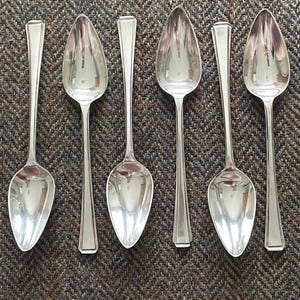 May include: Six silver spoons with a pointed oval bowl and a long, narrow handle. The spoons are arranged in a row on a brown and white patterned fabric.