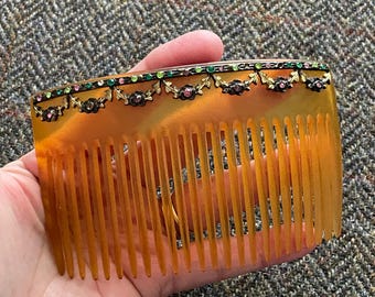 May include: A vintage tortoiseshell comb with a decorative row of rhinestones and green and pink stones. The comb is made of a dark brown material and has a curved shape.