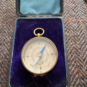 May include: A vintage brass compass with a blue and white face, resting in an open black case with a blue velvet interior. The compass needle points north. The case is rectangular with a gold-colored ring at the top.