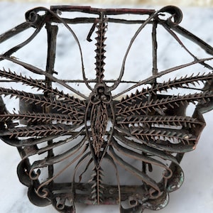May include: A vintage, ornate metal butterfly-shaped object with intricate details. The object is made of dark, aged metal with a rusty appearance. The butterfly's wings and body are clearly defined, showcasing a detailed design. The object is likely a decorative piece or a mold.