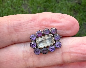 May include: A vintage ring featuring a rectangular, clear central stone surrounded by a halo of smaller, round, purple gemstones. The ring has a gold-colored setting and is displayed against a blurred green background.