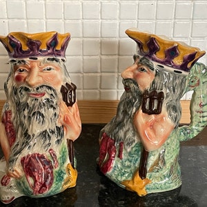 May include: Two ceramic jugs shaped like mermen with beards and crowns. The jugs are decorated with green, yellow, red, and brown paint. One jug has a seahorse handle.