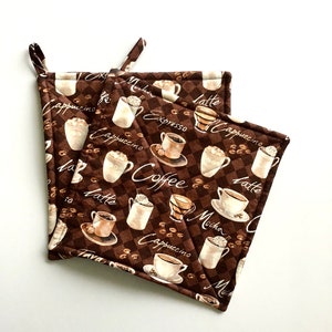 May include: Two brown quilted pot holders with a coffee theme. The pot holders feature a variety of coffee drinks, including cappuccino, latte, mocha, and espresso. The pot holders are decorated with a checkered pattern and coffee beans.