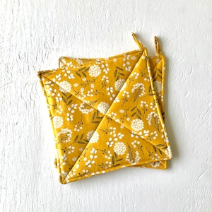 May include: Two yellow pot holders with a white floral pattern. The pot holders are folded in half and have a square shape.