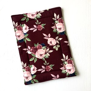 May include: A burgundy quilted fabric placemat with a floral pattern featuring pink and white roses and green leaves.