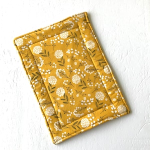May include: Mustard yellow rectangular padded mat with a floral pattern of white flowers and green leaves. The mat has a stitched border and a smaller, inset rectangle. The floral design is repeated across the surface.