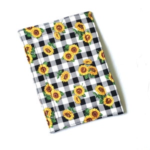 May include: A rectangular, padded item featuring a black and white checkered pattern. The pattern is overlaid with yellow sunflowers with green leaves. The item is likely a kitchen accessory, such as a pot holder or trivet.