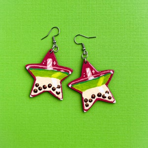 May include: A pair of star-shaped earrings with a unique design. The earrings feature a red border, green and white layers, and brown dots, resembling a beverage. The earrings hang from silver-colored hooks, set against a bright green background.