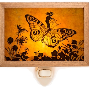 Puede incluir: Una luz nocturna con un marco de madera y un fondo amarillo. La imagen dentro del marco es una silueta de un hada montando una mariposa con flores en el fondo.