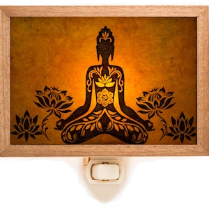 Peut inclure: Une veilleuse avec un cadre en bois et un insert en verre représentant une silhouette noire d'un Bouddha en méditation avec des motifs floraux complexes. Le Bouddha est entouré de fleurs de lotus et a un mandala jaune lumineux au centre de sa poitrine.