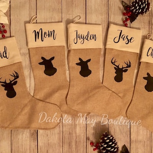 May include: Five burlap Christmas stockings with white fabric tops and deer silhouettes. The stockings are personalized with the names "Dad", "Mom", "Jayden", "Jase", and "Chelsey".