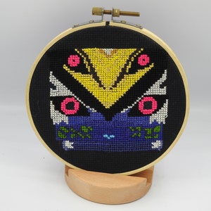May include: A black embroidery hoop with a colorful geometric design featuring yellow, pink, blue, white, and green. The design is a stylized butterfly with a yellow center and pink eyes.