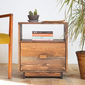 May include: A wooden nightstand with a drawer and a shelf. The nightstand is stained a dark brown color and has a rustic look. There are books on the shelf, including "The Good New Days" by  Judd  Calment.