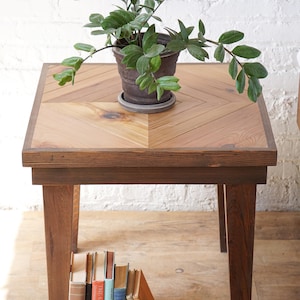 May include: A wooden side table with a herringbone pattern top, supporting a potted plant. The table has dark brown legs and a collection of books leaning against them. The plant has green leaves. The table is approximately 24 inches tall.