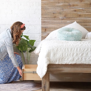 May include: A woman is kneeling down and opening a drawer on a wooden bed frame. The bed has a white comforter and a light blue pillow with white embroidery. The headboard is made of wood planks.