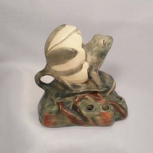 May include: A ceramic frog figurine with a green and white striped shell on its back. The frog is sitting on a green and brown base with holes for holding flowers.