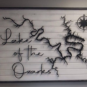 May include: A black and white wooden sign with the text "Lake of the Ozarks" in a cursive font. The sign also features a black metal outline of a lake with a compass rose in the top right corner.