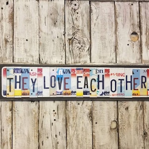 May include: A framed sign made of license plates spelling out the phrase "They Love Each Other". The sign is on a white wood background.