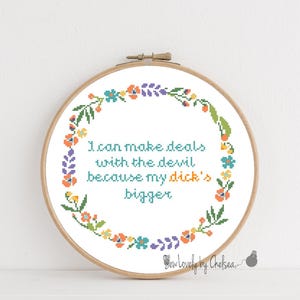 Puede incluir: Un bastidor de punto de cruz con el texto "I can make deals with the devil because my dick's bigger" en turquesa. El texto está rodeado por una corona floral en tonos naranja, azul, verde y morado.