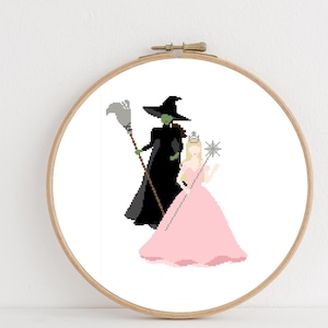 Puede incluir: Un bastidor de bordado de punto de cruz con imágenes pixeladas de la Malvada Bruja del Oeste y Glinda la Bruja Buena de El Mago de Oz. La bruja está en negro, sosteniendo una escoba, y Glinda está en rosa, sosteniendo una varita.