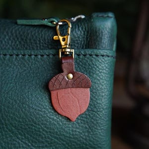 May include: A close-up of a dark green leather pouch with a brown and red acorn-shaped leather charm attached by a gold-colored clasp. The charm has a textured top and a smooth, rounded bottom.