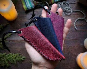 May include: Three leather pouches with drawstrings, one red, one dark blue, and one burgundy. The pouches are held in a hand against a wooden background.