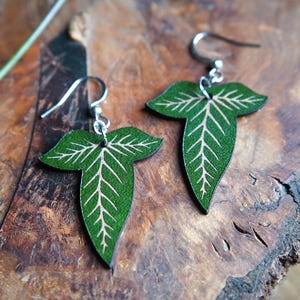 May include: Green leaf-shaped earrings with white vein detailing, suspended from silver hooks. The earrings are presented on a textured, brown wooden surface, with a blurred background of wood and foliage.
