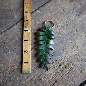 A green leather fern keychain is laying on a rustic wooden table next to an antique folding wood ruler. It shows the measurement to be around 5 inches in length.