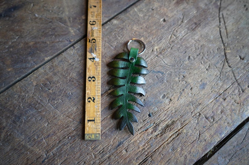 A green leather fern keychain is laying on a rustic wooden table next to an antique folding wood ruler. It shows the measurement to be around 5 inches in length.