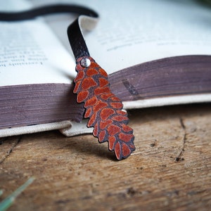 May include: A brown leather bookmark shaped like a pine cone with a black leather strap. The bookmark is lying on a book.