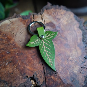 May include: A green leather leaf-shaped keyring with a silver ring and a silver rivet. The keyring is on a wooden surface.
