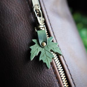 May include: A dark brown textured leather bag featuring a silver metal zipper closure. Attached to the zipper pull is a green leather maple leaf charm secured with a small brass-colored rivet, adding a distinct detail to the bag's exterior.