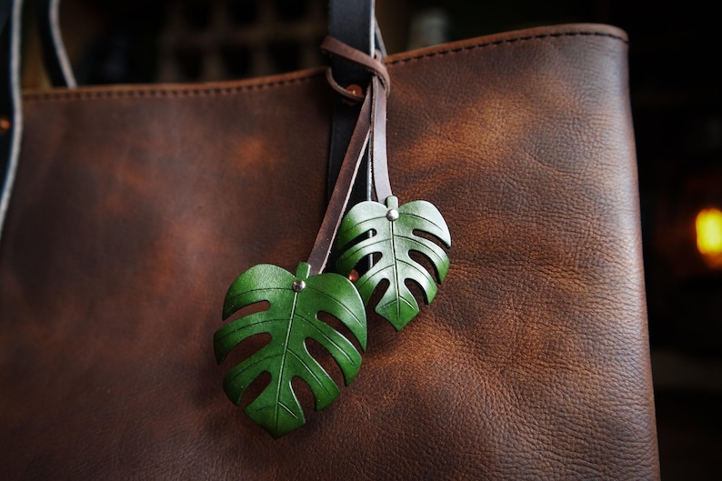 A leather bag charm with two monstera leaves in a deep green, riveted to a brown strap that is wrapped around the handle of a rich brown pebbled leather tote bag. There is a lantern glowing in the background