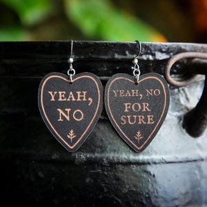 May include: Two heart-shaped earrings with the phrases "YEAH, NO" and "YEAH, NO FOR SURE" in copper-coloured text. The earrings are black with a copper-coloured border and a small leaf design. They hang from silver-coloured hooks.