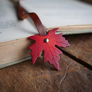 May include: A red leather maple leaf bookmark with a brown leather strap, resting on the pages of an open book. The bookmark is secured with a silver rivet. The book is on a wooden surface.