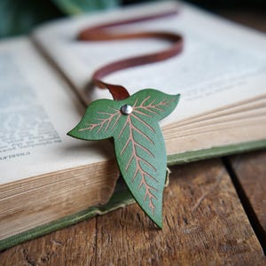 May include: A green leather bookmark shaped like a leaf with a brown leather strap. The leaf has a detailed vein pattern and a silver rivet.