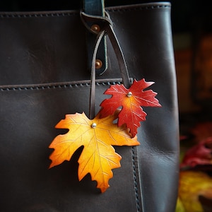 May include: A brown leather tote bag with a leather strap and two leather maple leaf charms. The charms are orange and red.