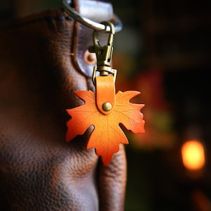May include: A leather keychain featuring a maple leaf design in shades of orange and yellow, attached to a bronze-colored clasp. The keychain is connected to a brown leather bag, with a blurred background.