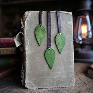 May include: Three green leather leaf bookmarks with brown leather straps hanging from a closed book with a tan cover.