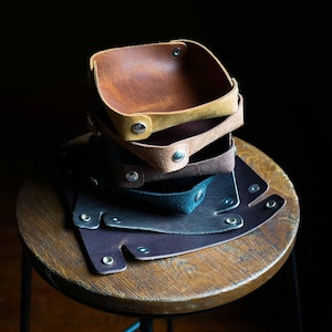 May include: A stack of leather catchall trays in various colors, including brown, tan, and blue. Each tray is square with rounded corners and secured with metal rivets. The trays are displayed on a wooden stool against a dark background.