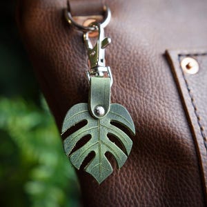 May include: A sage green leather keychain shaped like a monstera leaf, featuring a silver-toned metal lobster clasp and a central silver rivet. The leaf design includes cut-out details and visible veining, attached to a brown leather bag.