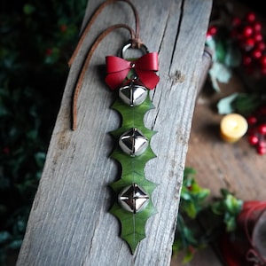 May include: A green leather holly leaf ornament with four silver bells and a red bow. The ornament is hanging from a brown leather cord.