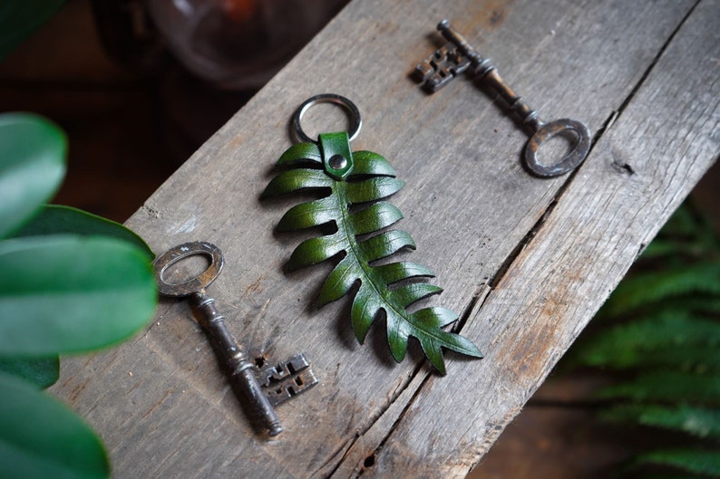 A molded 3D effect green leather fern keychain is sitting on a rustic wood board with two antique skeleton keys sitting next to it. A real fern is blurred in the background.
