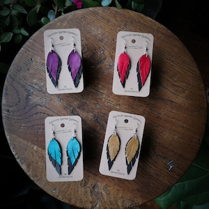 May include: Four pairs of feather-shaped earrings made from recycled leather. Each pair features a different color combination: purple and black, red and black, teal and black, and gold and black. The earrings are displayed on a wooden surface with a card that reads "Recycled Leather Earrings" and "Everyday Well Made".